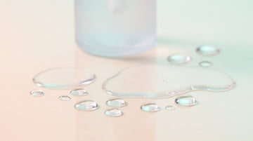 Clear lubricant droplets and a frosted glass bottle on a soft pastel surface — personal lube guide